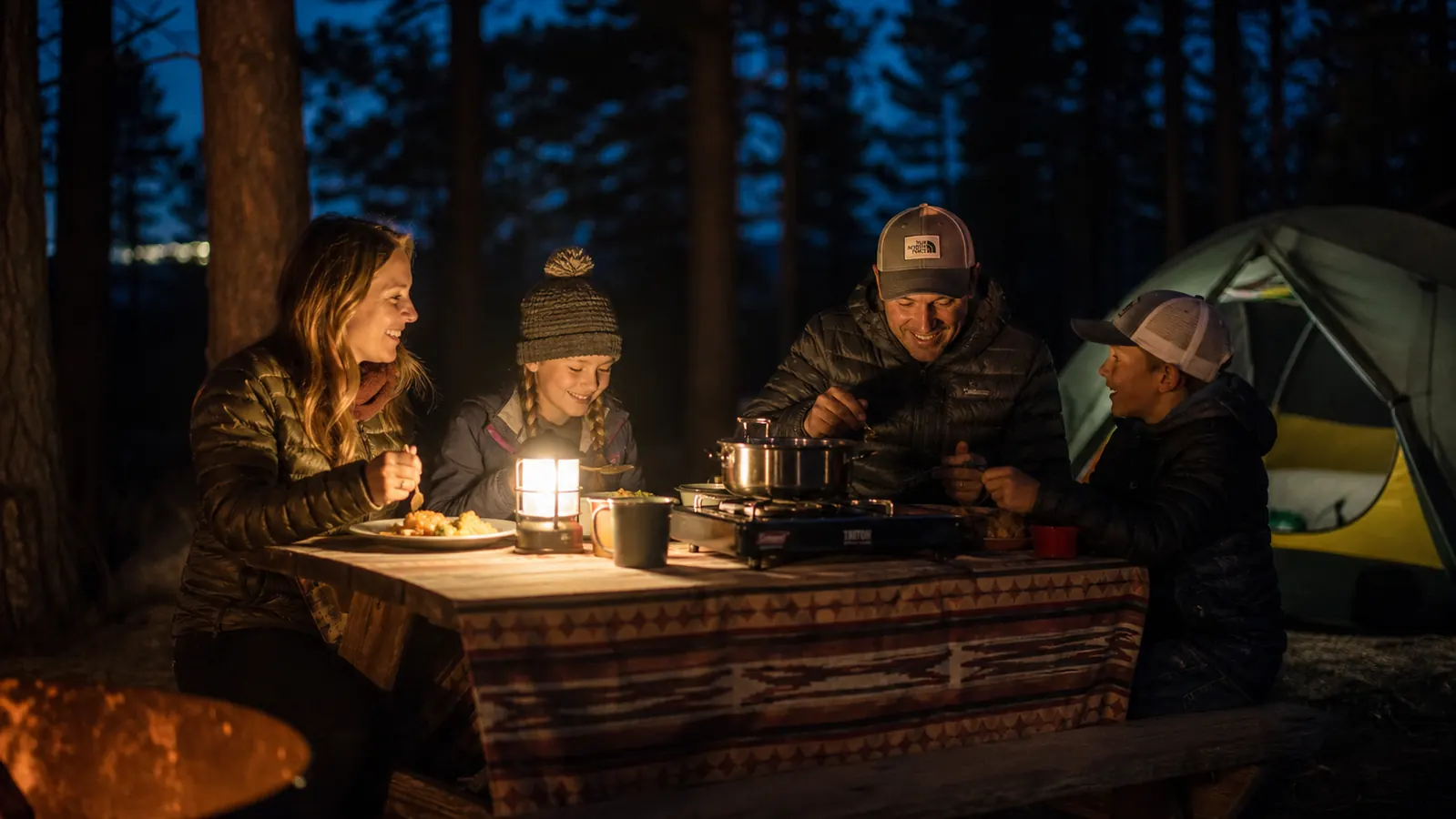 Camper eating a hot meal beside a tent as the sun sets over a ridgeline