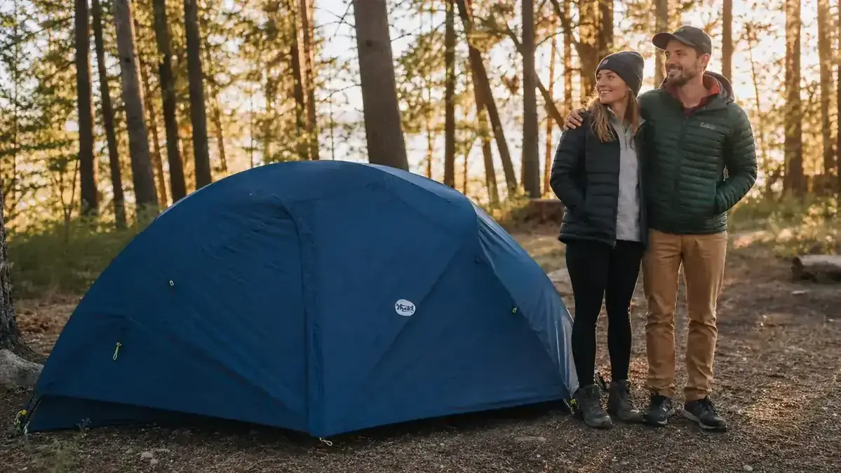 Camper sitting in open tent doorway with coffee, morning light, forest behind
